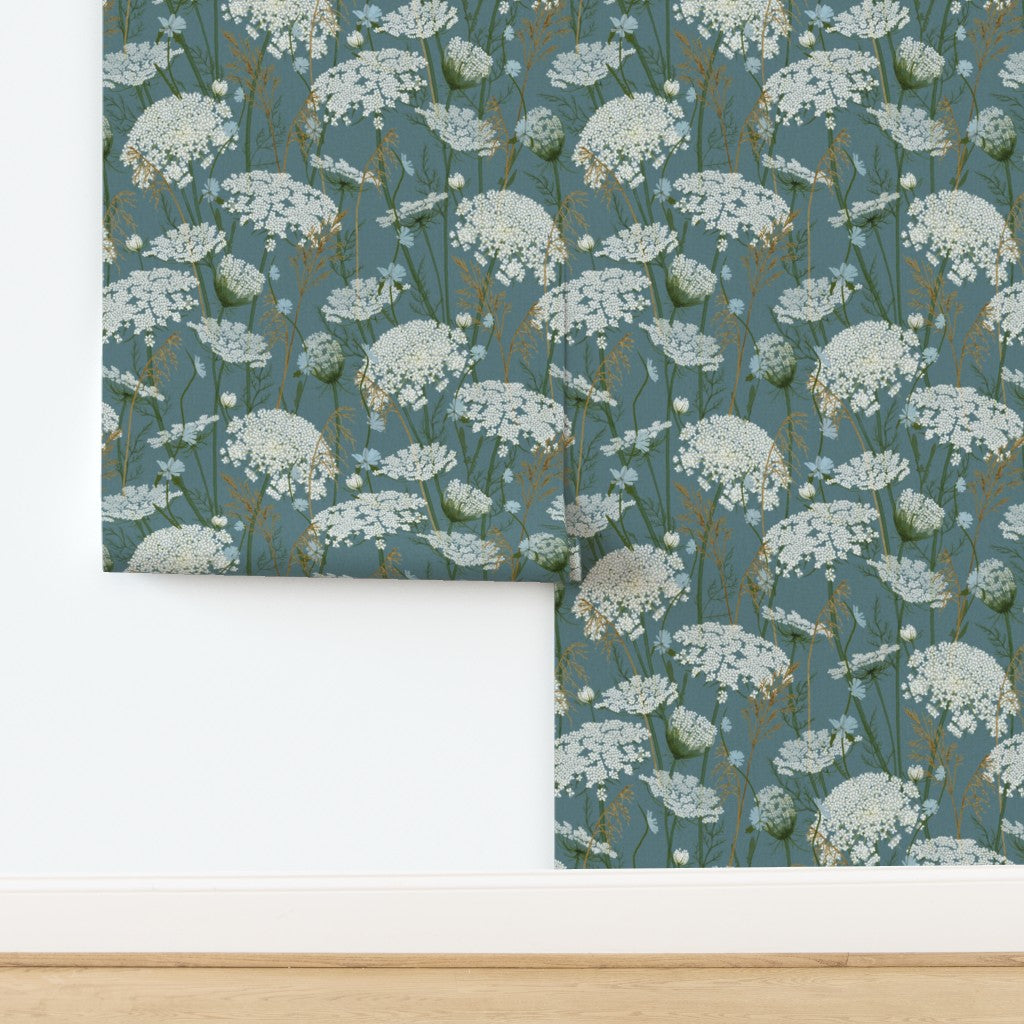 A folded piece of wallpaper with a repeating white wildflower pattern on a muted blue-gray background, displayed against a white wall above a light wood floor.